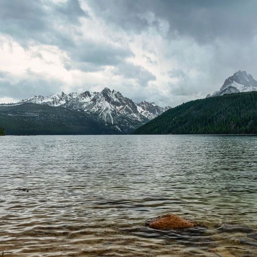 A gorgeous landscape with tall snowy peaks and forests surrounding a calm lake under a cloudy sky