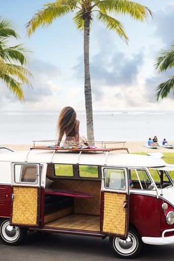 A vertical shot of a Caucasian female sitting on a van parked on a beach against palm trees and a sea