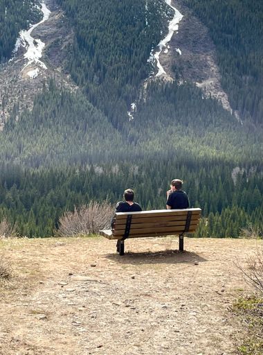 A back view of young boys sitting on bench in front of mountains surrounded by dense trees