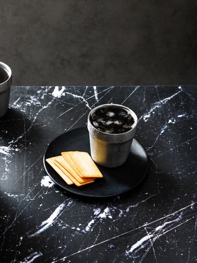 A vertical shot of crackers in a black plate and a steel cup with ice on a granite surface table