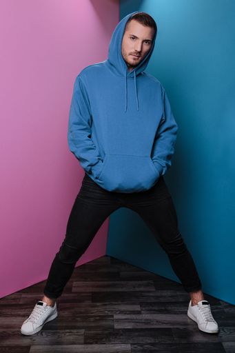 A vertical shot of a man in a blue hoodie standing legs apart and his arms in pockets on pink-blue background