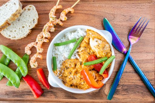 A top shot of a rice dish in a white bowl, grilled shrimp skewers with bread on a wooden board
