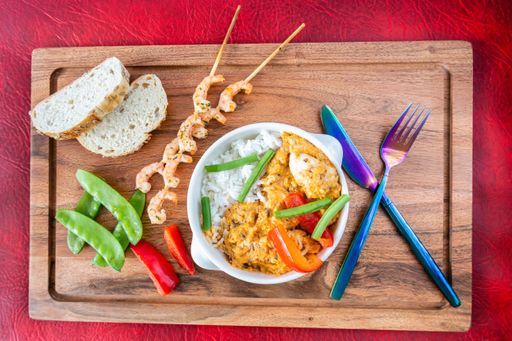 A top shot of a rice dish in a white bowl, grilled shrimp skewers with bread on a wooden board