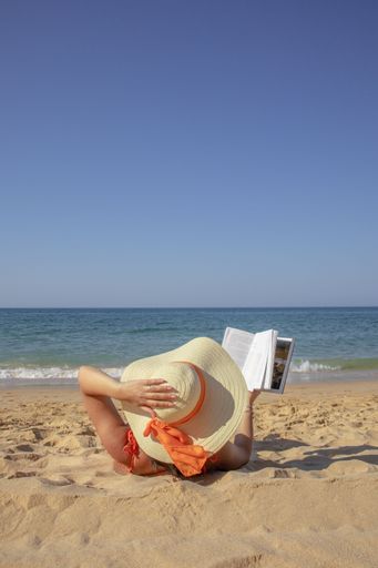 A rear view of a young girl in hat lying on sandy beach in Portugal and reading book