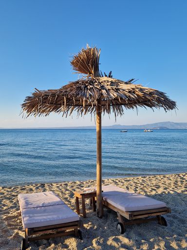 A vertical shot of two deck chairs on the beach under a straw umbrella with the sea and boats in the background.