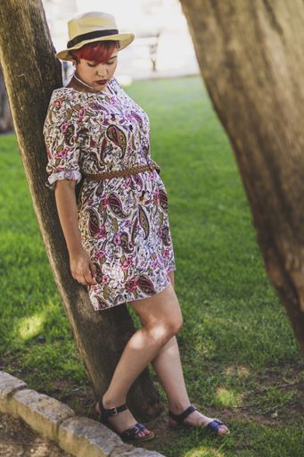 A vertical shot of a woman with red hair and hat leaning on the tree