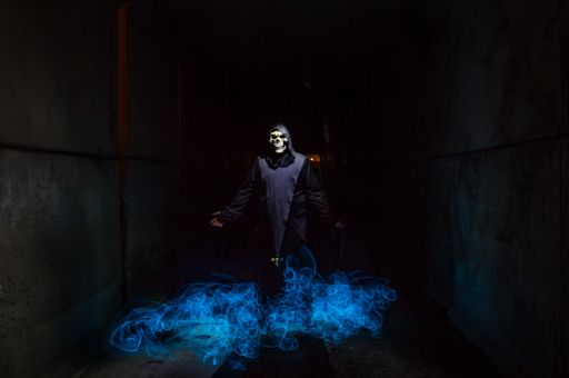 A person with a skull mask and black clothes with blue magical light - the concept of horror