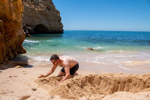 A shirtless Caucasian man building something with sand on the beach