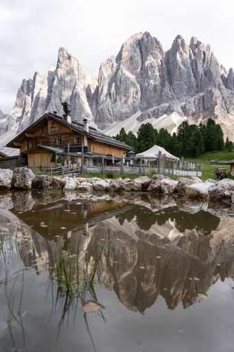 A vertical mesmerizing view of the rocky Geisler Alm mountains with a rural wooden house