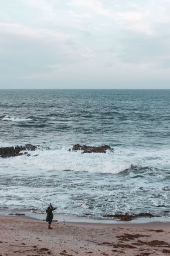 A vertical shot of a person standing on a sandy beach against ocean waves on a cloudy day