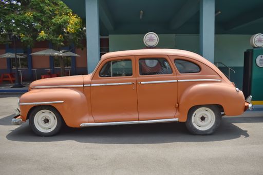 An Old Fashioned American Orange colored car resembling Plymouth De Luxe near a petrol Bunk against a green background. It is a vintage Car.
