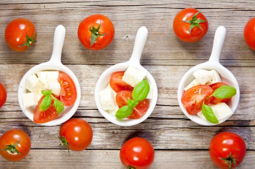 A top view of fresh mozzarella, tomato, and basil leaves in white bowls surrounded by whole tomatoes on a wooden surface