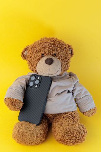A vertical view of the teddy bear wearing a beige top holding the smartphone on the green background
