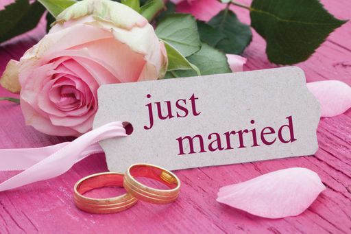 A "just married" tag with two golden wedding rings and a pink rose on a pink wooden surface
