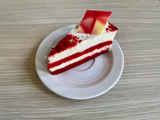 A piece of red velvet cake decorated with strip of white chocolate on a white plate lying on a wooden table.