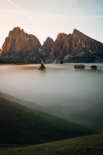 A vertical shot of rural houses and rocky mountains during a foggy sunrise in Seiser Alm, South Tyrol