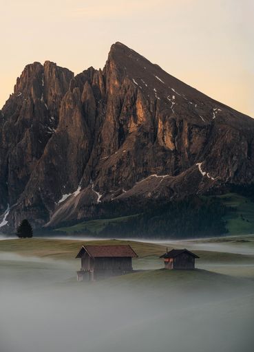 A vertical shot of rural houses and rocky mountains during a foggy sunrise in Seiser Alm, South Tyrol
