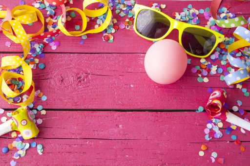 A top view of yellow sunglasses, balloon, confetti and birthday whistle on a pink wooden surface with space in the middle for text