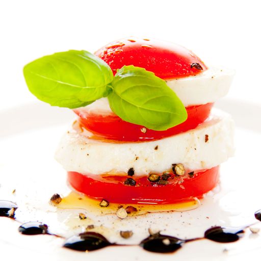 A closeup of fresh mozzarella cheese and slices of tomatoes assembled with basil, on a white plate decorated with balsamic vinegar