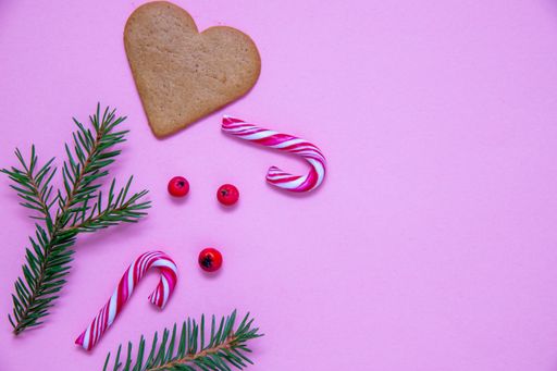 A flat lay shot of candy canes, fir branches, Christmas berries and a heart cookie on a pink surface