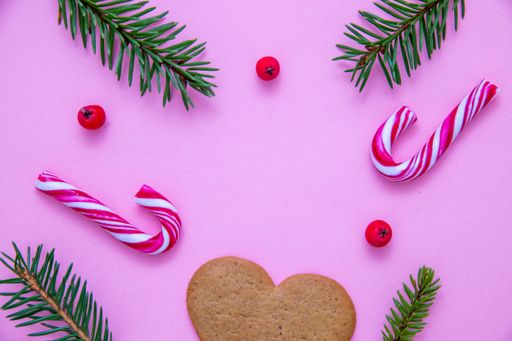 A flat lay shot of candy canes, fir branches, Christmas berries and a heart cookie on a pink surface