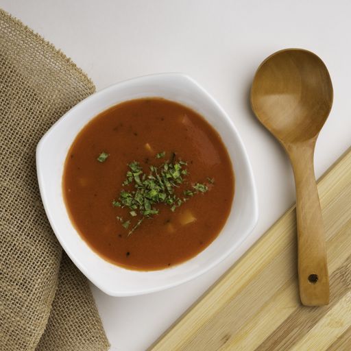 A high angle closeup shot of tomato soup with fresh parsley next to a wooden spoon and a napkin on a white table