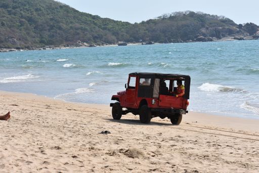 A red jeep car driving on a beach near a wavy sea with a hill in the background