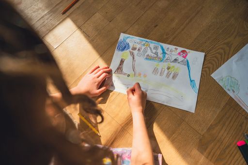 A high angle shot of a child drawing on a piece of paper on a wooden surface