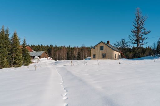 A wooden house in a snowy field with trees in the background during winter against a blue sky