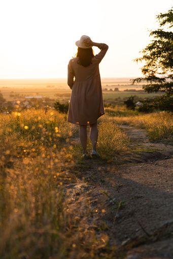 a back view of a cute girl with a dress and hat in a field during golden hour