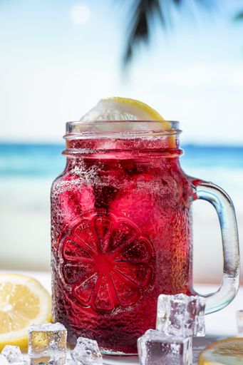 A vertical closeup shot of a red summer drink in transparent glass, ice and yellow lemons with a topical beach background