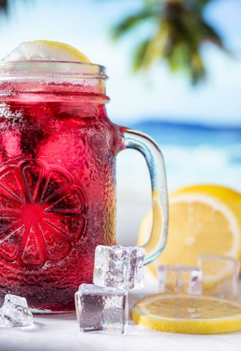 A vertical closeup shot of a red drink in transparent glass, ice, and yellow lemons with blur beach background