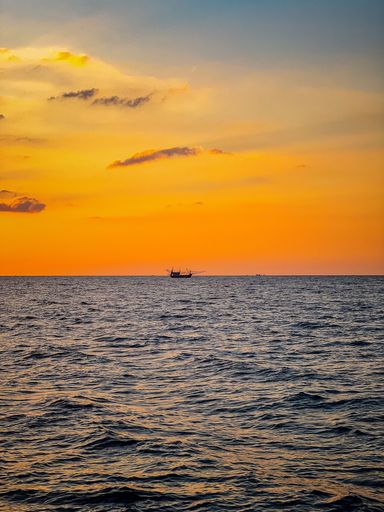 A vertical view of the silhouette of a boat on the horizon of seascape under the orange sunset sky