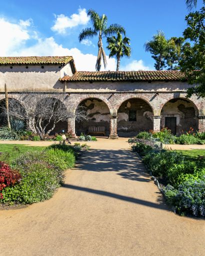 Brick arches and corridor taken from the garden at Mission San Juan Capistrano, with blue skies and palm trees, in San Juan Capistrano, California.