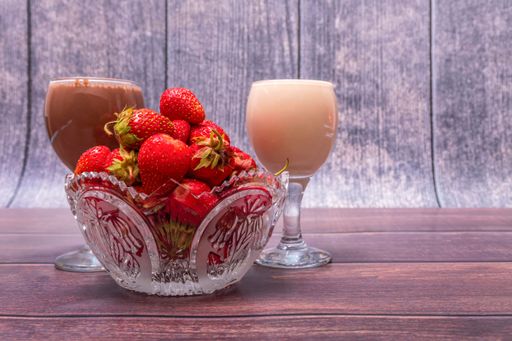 Crystal vase full of ripe red strawberries stands on wooden table. Nearby are wine glasses filled with milk and chocolate cocktails.