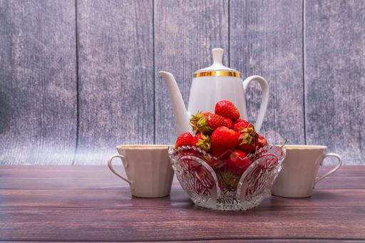 Glass vase filled with ripe red strawberries stands on wooden table. Nearby is porcelain teapot and two cups.