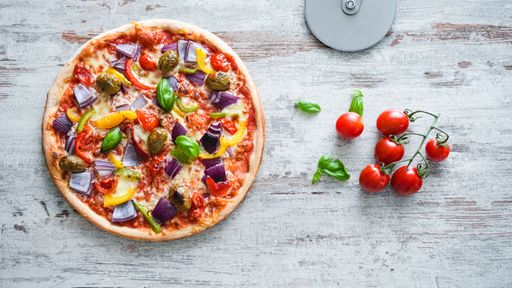 The top view of a mixed veggies pizza with cherry tomatoes and basil leaves over the gray wooden surface