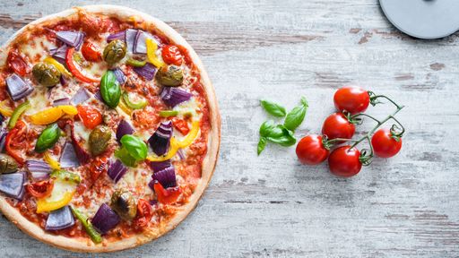 The top view of a mixed veggies pizza with cherry tomatoes and basil leaves over the gray wooden surface