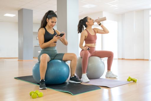 TWO GIRLS DOING PILATES AND YOGA, WHILE ONE IS ON THE PHONE AND ANOTHER DRINKS WATER rey happy