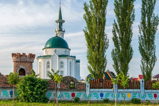Muslim mosque with high minaret and madrasah stands against of summer blue cloudy sky at sunset. In foreground, there is fence and pyramidal poplars.
