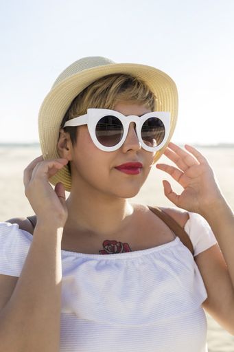 A vertical shot of a Caucasian female with short hair and a tattoo wearing sunglasses and a hat on the beach