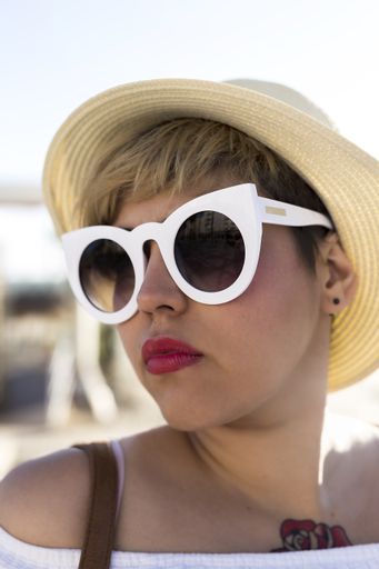 A vertical shot of a Caucasian female with short hair and a tattoo wearing sunglasses and a hat on the beach