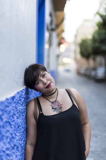 A vertical shot of a Caucasian female with short hair and a tattoo leaning against a wall