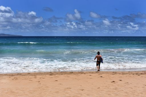 A back view of a man with surfboard on the sandy beach approaching the sea