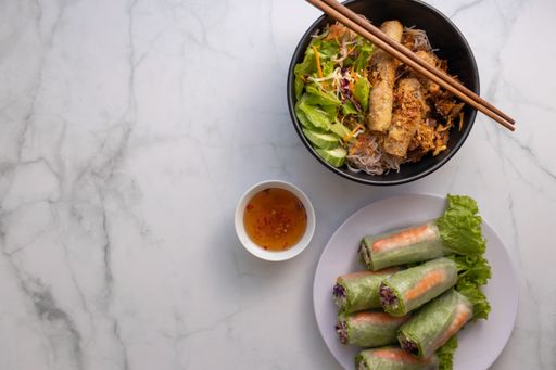 An overhead shot of a bowl of Vietnamese grilled pork on noodles, wooden chopsticks, fish sauce dip and fresh spring rolls with space for your text