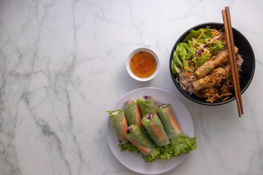 An overhead shot of a bowl of Vietnamese grilled pork on noodles, wooden chopsticks, fish sauce dip and fresh spring rolls and space for your text