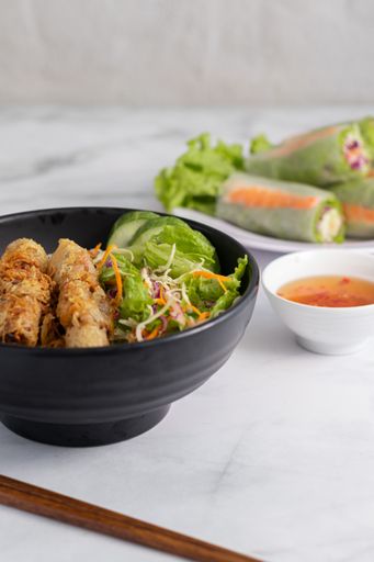 A vertical shot of a bowl of Vietnamese grilled pork on noodles with fish sauce dip and fresh spring rolls in the background