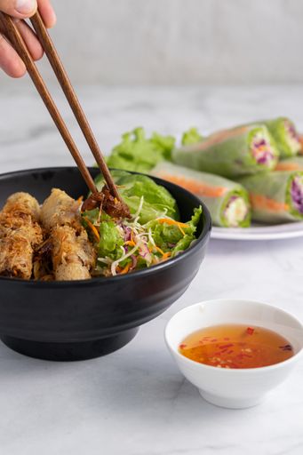 A vertical shot of a person eating Vietnamese grilled pork on noodles using chopsticks, with fish sauce dip and fresh spring rolls in the background