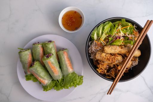 An overhead shot of a bowl of Vietnamese grilled pork on noodles, wooden chopsticks, fish sauce dip and fresh spring rolls on a white table