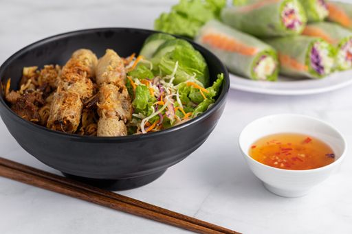A soft focus of a bowl of Vietnamese grilled pork on noodles with fish sauce dip and fresh spring rolls in the background